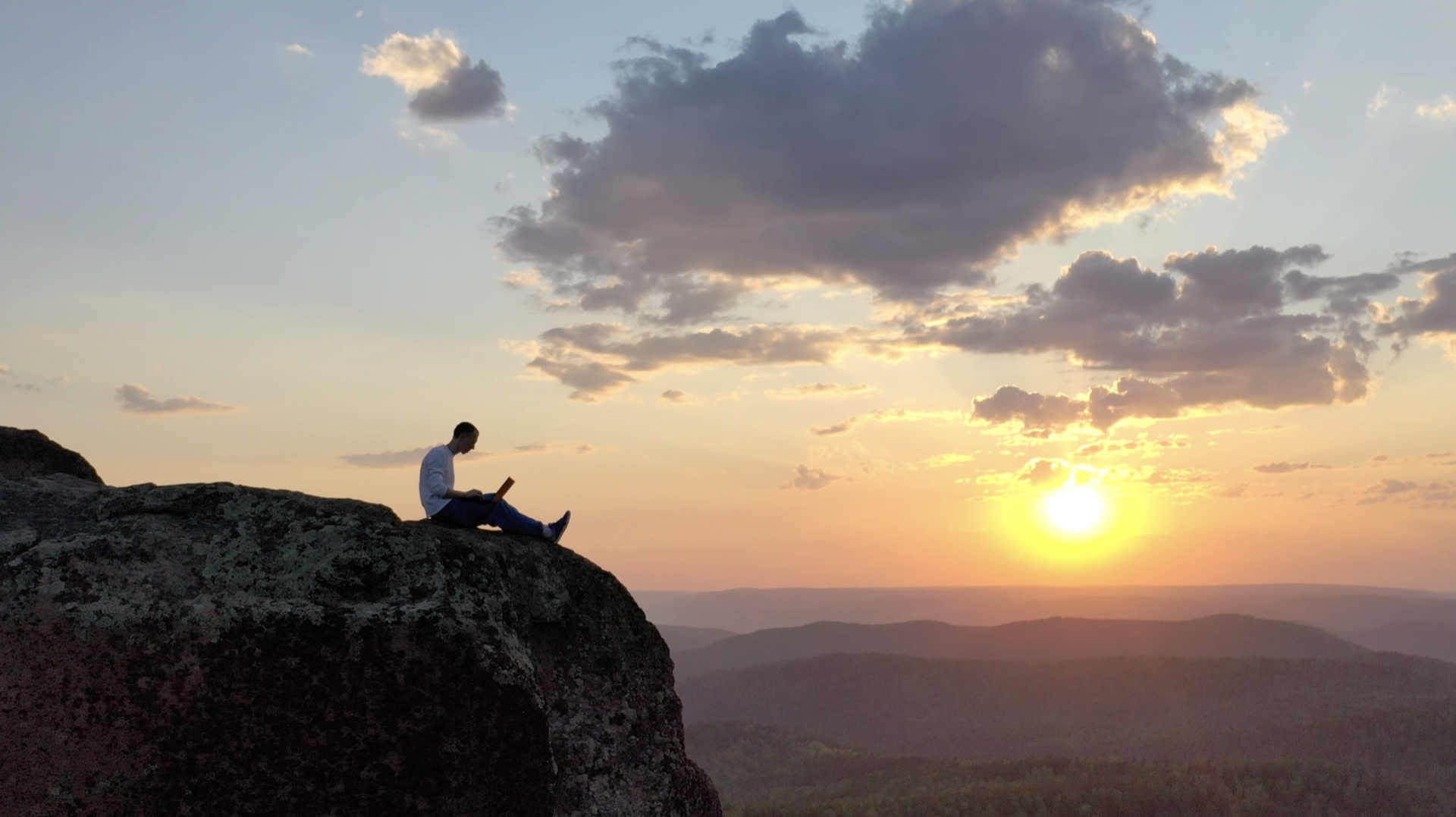 夕焼けの山を望む崖の上で、ノートパソコンを開いて仕事をする人のシルエット
